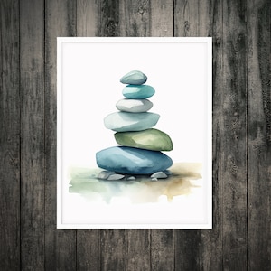 Puede incluir: Una impresión en acuarela enmarcada que presenta una pila de piedras lisas y redondeadas en tonos azules y verdes. Las piedras están equilibradas, creando una composición zen. La obra de arte está sobre un fondo blanco.