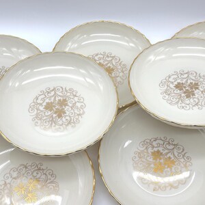 May include: A collection of off-white ceramic bowls with a gold floral design in the center. The bowls have a scalloped edge and a gold rim. The bowls are arranged in a circular pattern.