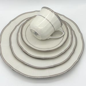 May include: A white porcelain tea set with a silver trim. The set includes a teacup, saucer, and three plates of varying sizes. The plates and teacup have a ridged pattern. The edges of the plates have a scalloped design.