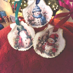 May include: Three seashell Christmas ornaments, each featuring a snowman design. The snowmen wear hats and scarves in various colors, surrounded by floral accents. Gold trim edges the shells, and ribbons are attached for hanging.