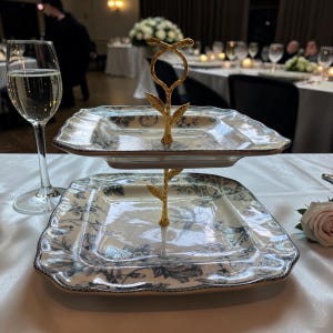 May include: Two-tiered serving tray with a gold-colored metal stand. The square plates have a blue and white floral design. The tray is set on a white tablecloth, with a glass of champagne and a rose in the background.