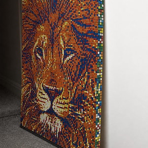 May include: A rectangular wall art piece constructed from a grid of small, multi-colored square tiles. The mosaic depicts a lion's face in shades of orange, blue, yellow, and white. The side edge shows a vertical column of various colored squares.