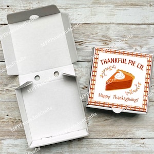 Può includere: Una scatola per pizza in cartone bianco con un inserto a tema del Ringraziamento. L'inserto presenta uno sfondo a scacchi rosso e bianco con una fetta di torta di zucca e il testo "THANKFUL PIE CO. Happy Thanksgiving!"