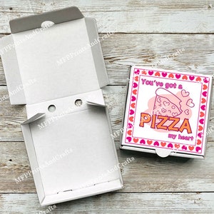 May include: White pizza box with the text "You've got a PIZZA my heart" in pink and orange. The box has a pizza slice graphic with hearts. The box is open, revealing the inside.