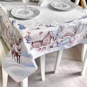 May include: A rectangular tablecloth with a winter scene design featuring a cabin, bridge, and deer. The tablecloth is set on a white table with white chairs, plates, and wine glasses. The color palette includes white, blue, and brown.