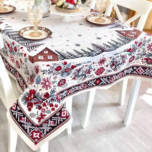 Può includere: Una tovaglia bianca con un design natalizio festivo con motivi floreali e geometrici rossi e neri e un'illustrazione di una baita. Il tavolo è apparecchiato con sedie bianche, bicchieri e piatti.