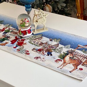 Op de afbeelding: Een rechthoekige tafelloper met een kerstthema, met een winterse scène met rendieren, sneeuwpoppen en kinderen die sleeën. De loper is versierd met een sneeuwbol en een decoratief hertfiguur. De tafelloper is ongeveer 150 cm lang.
