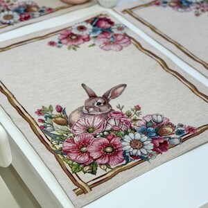 May include: A set of four rectangular placemats with a floral and bunny design. Each placemat features a beige background with a brown border and a detailed illustration of a bunny surrounded by pink, blue, and white flowers. A pink bunny figurine and a small vase with flowers are on the table.