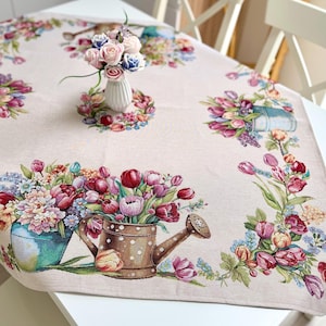 Può includere: Tovaglia floreale beige con un colorato design di tulipani e fiori. La tovaglia presenta immagini di annaffiatoi e vasi pieni di fiori. Un piccolo vaso di fiori rosa e blu è al centro del tavolo.