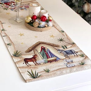 Può includere: Runner da tavolo natalizio beige con una scena della Natività, raffigurante Maria, Giuseppe e il bambino Gesù in una mangiatoia. Il runner è decorato con stelle e verde. Un candelabro dorato e un centrotavola decorativo con candele e ornamenti sono sul tavolo.