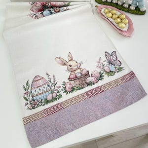 Könnte beinhalten: Weißer Tischläufer zum Thema Ostern mit lavendelfarbenem Rand. Der Läufer zeigt Illustrationen von Ostereiern, Hasen und Schmetterlingen. Eine dekorative Hasenfigur und eine Schale mit goldverpackten Süßigkeiten befinden sich ebenfalls auf dem Tisch.