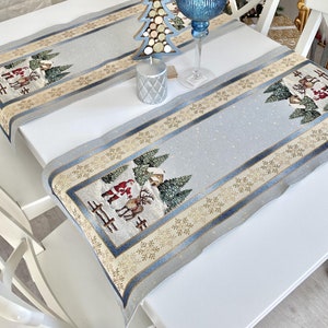 May include: Two light blue Christmas table runners with a festive design featuring Santa Claus, reindeer, and snowy trees. The runners have a gold and blue border and are placed on a white table, creating a holiday atmosphere.