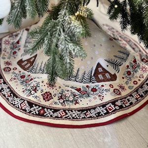 May include: A beige Christmas tree skirt with a red border and a winter village scene. The skirt features houses, trees, and snowflakes. The skirt is under a decorated Christmas tree.