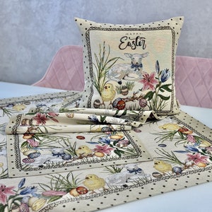 May include: A set of three beige table runners with a black polka dot pattern. The runners feature an embroidered Easter design with chicks, lambs, flowers, and eggs. The design is in shades of pink, yellow, green, and blue. The runners are bordered with a black and white trim. The center runner is folded in half. The runners are on a white table with a pink chair in the background. The pillow in the background has a white lamb and the text "Happy Easter".