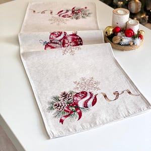 May include: Beige Christmas table runner featuring embroidered red baubles, snowflakes, pine cones, and ribbons. The rectangular runner is made of textured fabric, ideal for festive decorations.