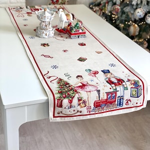 Può includere: Runner da tavolo natalizio color crema con bordo rosso. Il runner presenta illustrazioni festive di una ballerina, schiaccianoci, albero di Natale e trenino giocattolo. Oggetti decorativi come una giostra e una palla di neve sono sul tavolo.