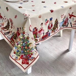 Op de afbeelding: Een crèmekleurig kersttafelkleed met een feestelijk Notenkraker- en kerstboomontwerp. De rand is rood en de stof is versierd met vakantie-thema illustraties, waaronder cadeaus, ornamenten en sneeuwvlokken. Het tafelkleed ligt op een witte tafel met borden en glazen.
