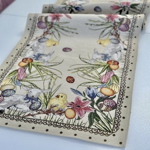 Può includere: Runner da tavolo beige a tema pasquale con un design floreale e animale dettagliato. Presenta agnelli, pulcini, uova di Pasqua e fiori dai colori pastello. I bordi sono rifiniti con un bordo a pois e un motivo in pizzo decorativo.
