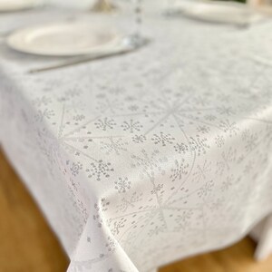Può includere: Tovaglia bianca con motivo a fiocchi di neve. La tavola è apparecchiata con piatti e bicchieri, con un albero decorativo sullo sfondo. Il motivo a fiocco di neve è di colore argento chiaro.