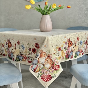 May include: A square, cream-colored tablecloth with a colorful Easter design. The tablecloth features bunnies, chicks, flowers, and eggs. The tablecloth is laid on a table with four blue chairs.