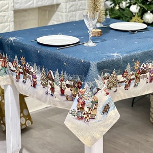 Op de afbeelding: Een blauw kerstkleed met een winterdorpsscène. De rand heeft een gedetailleerde illustratie van mensen, huizen en bomen in een besneeuwd landschap. Twee witte borden en een glas staan op de tafel.