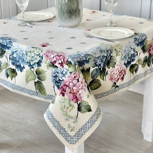 Pode incluir: Uma toalha de mesa branca com um padrão floral com hortênsias rosa, azuis e verdes. A toalha de mesa está posta sobre uma mesa branca com dois talheres e um vaso de hortênsias no centro.