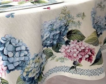 以下が含まれることがあります： 青とピンクのヒアシンスの花柄が描かれた白いテーブルクロス。テーブルクロスは、幾何学模様の青と白の縁取りが施されています。