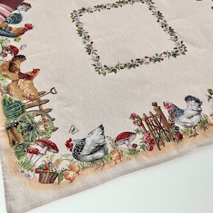 May include: Beige rectangular tablecloth with a detailed farm scene border. The border features chickens, mushrooms, flowers, and a wooden fence. A square floral design is in the center. The tablecloth is on a white surface.
