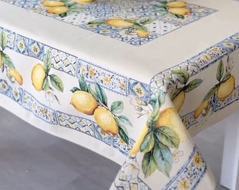 May include: A cream-colored tablecloth with a lemon and leaf pattern, a matching pillow, and a yellow pot with a green plant. The tablecloth has a blue and yellow border with a repeating geometric design. The pillow has a lemon and leaf design.