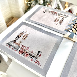 Op de afbeelding: Twee grijze placemats met een witte achtergrond en een grijze rand. Een placemat heeft een vintage trein met een rode locomotief en een notenkraker. De andere placemat heeft een notenkraker en een speelgoedtrein.