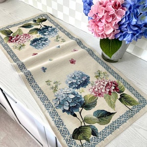 May include: A beige rectangular table runner with a floral hydrangea design. The runner features blue, pink, and green hydrangeas with green leaves. A blue geometric border frames the floral pattern. Butterflies are scattered across the runner.