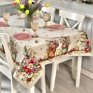 Könnte beinhalten: Eine weiße Tischdecke mit einem floralen Muster und Ostereiern. Die Tischdecke hat einen Rand aus bunten Blumen, Eiern und Körben. Die Tischdecke liegt auf einem Tisch mit vier weißen Stühlen.