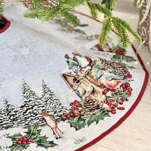 May include: A white and blue Christmas tree skirt with a red border. The skirt features a winter scene with a birdhouse, birds, holly, berries, and a deer.