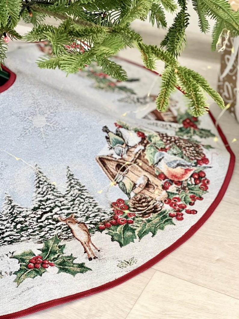 May include: A white and blue Christmas tree skirt with a red border. The skirt features a winter scene with a birdhouse, birds, holly, berries, and a deer.
