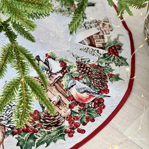 May include: A white and red Christmas tree skirt with a winter scene featuring a bird, pine cones, berries, and a small house. The skirt has a red border.