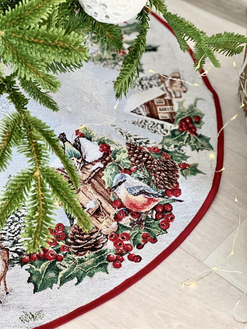May include: A white and red Christmas tree skirt with a winter scene featuring a bird, pine cones, berries, and a small house. The skirt has a red border.