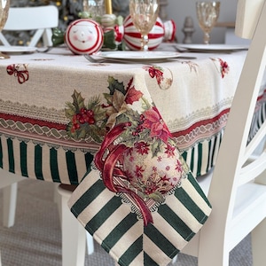 Peut inclure: Une nappe rectangulaire avec un centre couleur crème orné d'un motif festif de rubans rouges, de baies de houx et de poinsettias. La bordure présente une large bande de rayures verticales vert foncé et crème avec des détails en dentelle.