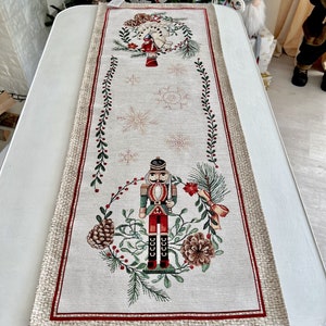 May include: A beige table runner with a red border features a festive Christmas design. The center of the runner depicts a nutcracker soldier standing in a wreath of greenery with pine cones and red berries. The design is repeated at the top of the runner with a smaller version of the nutcracker.