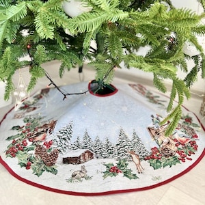 May include: A white and blue Christmas tree skirt with a winter scene design. The skirt features a snowy forest with trees, a cabin, a deer, and a rabbit. The skirt has a red border.