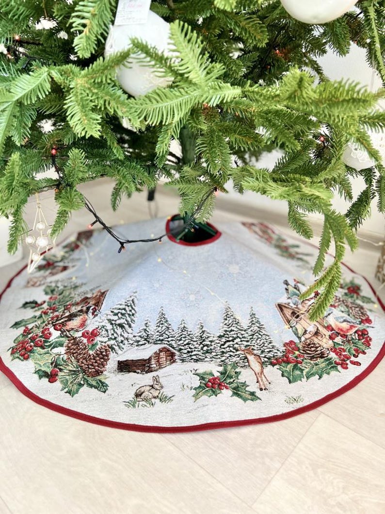 May include: A white and blue Christmas tree skirt with a winter scene design. The skirt features a snowy forest with trees, a cabin, a deer, and a rabbit. The skirt has a red border.