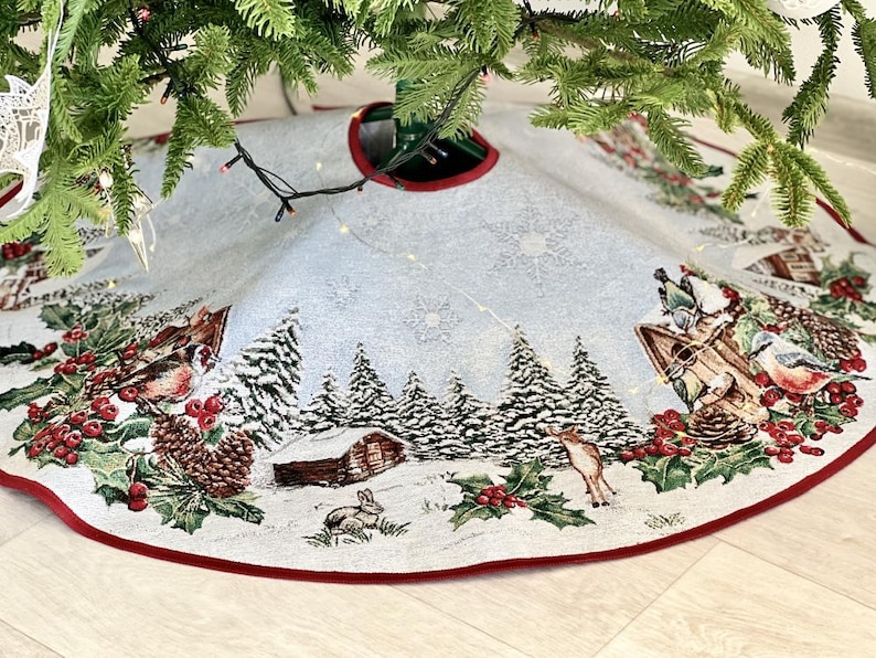 May include: A white and red Christmas tree skirt with a winter scene featuring a cabin, trees, birds, and a deer. The skirt has a red border and a white background with a snowflake pattern.