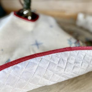 May include: White quilted Christmas tree skirt with a red velvet trim. The skirt has a star pattern.