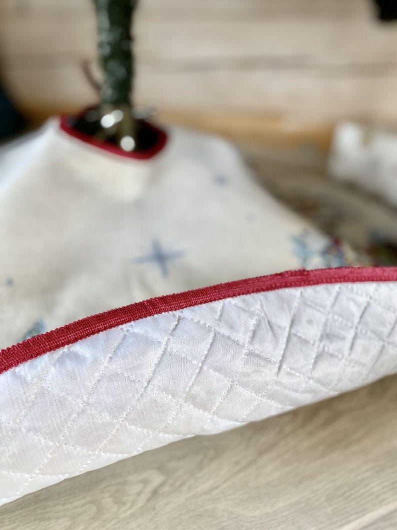 May include: White quilted Christmas tree skirt with a red velvet trim. The skirt has a star pattern.