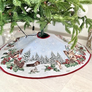 May include: A white and red Christmas tree skirt with a winter scene design. The skirt features a snowy forest with trees, houses, birds, and a deer. The skirt has a red border.