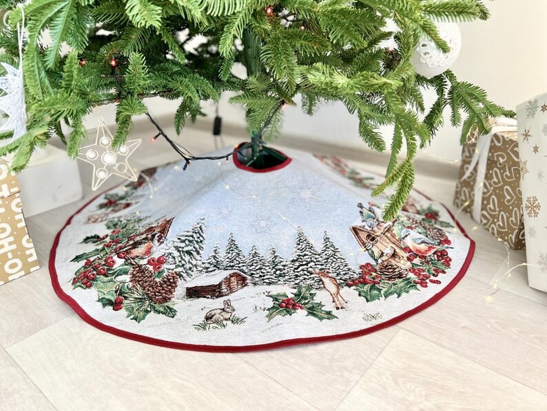 May include: A white and red Christmas tree skirt with a winter scene design. The skirt features a snowy forest with trees, houses, birds, and a deer. The skirt has a red border.