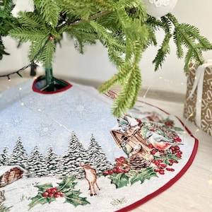 May include: A white and red Christmas tree skirt with a winter scene featuring a cabin, trees, a deer, and birds. The skirt has a red border and a white background.