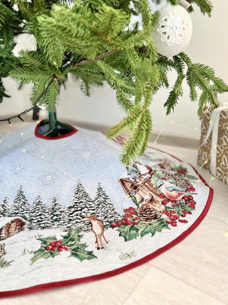 May include: A white and red Christmas tree skirt with a winter scene featuring a cabin, trees, a deer, and birds. The skirt has a red border and a white background.