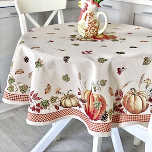 Op de afbeelding: Een ronde tafelkleed met een herfstthema. De crèmekleurige stof heeft pompoenen, bladeren en bessen in tinten oranje, bruin en groen. Een rood-bruine geblokte rand omlijst het ontwerp. Een decoratieve haan kan bloemen bevatten.