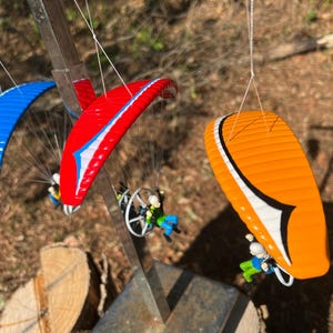 May include: Three miniature paragliders in blue, red, and orange hang from a metal structure. Each paraglider has a pilot figure attached. The paragliders are suspended against a blurred outdoor background.