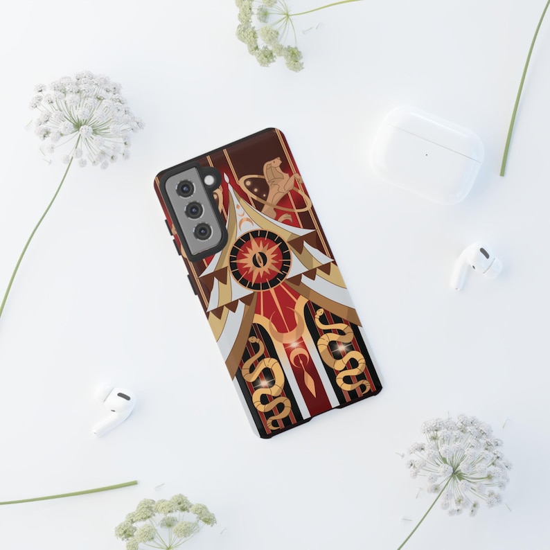 May include: A phone case with a detailed design in shades of red, brown, gold, and white. The case features a central sunburst design, geometric shapes, and stylized snakes. The phone case is on a white surface with white flowers and earbuds.